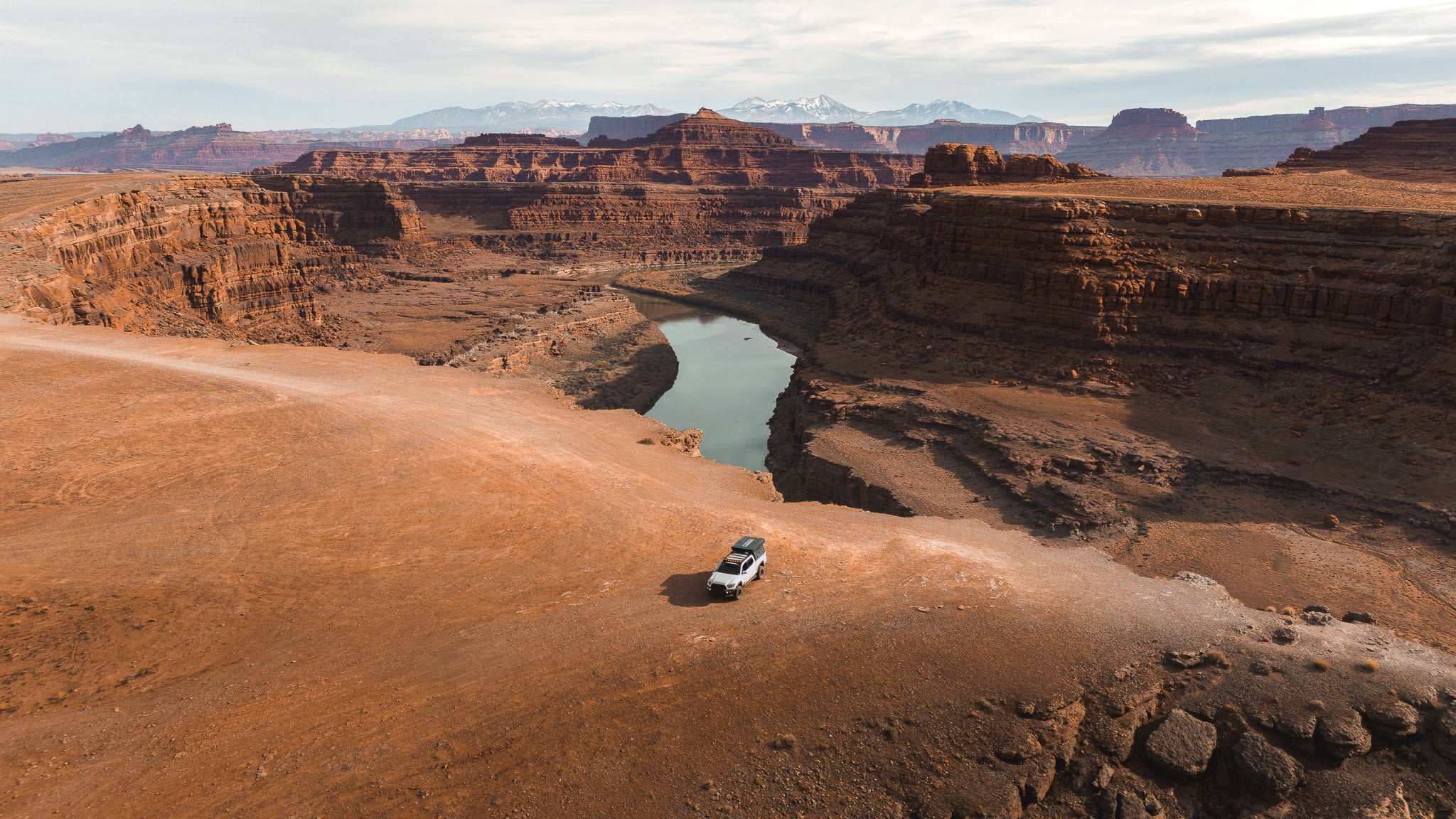 An overland truck with a rooftop tent parked on a high desert canyon overlook above a winding river, showcasing the extended range capability of Helivue Design auxiliary fuel systems.